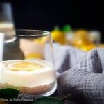 Lemon cheesecake layered in glass on top of biscuit layer. Lemons and grey blue cloth blur in background against a black backdrop