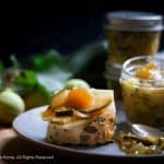 Green apricot relish on top of slices of cheese, served on top of a slice of seedy bread. The topped bread sits on a speckled grey blue place, an opened jar of the green apricot relish sits on the plate next to a smear of the relish in which a silver teaspoon lay. In the background is fresh uncooked whole green apricots with some leaves from an apricot tree. unopened jars of the green apricot relish can be seen slightly out of focus blurred in the background.